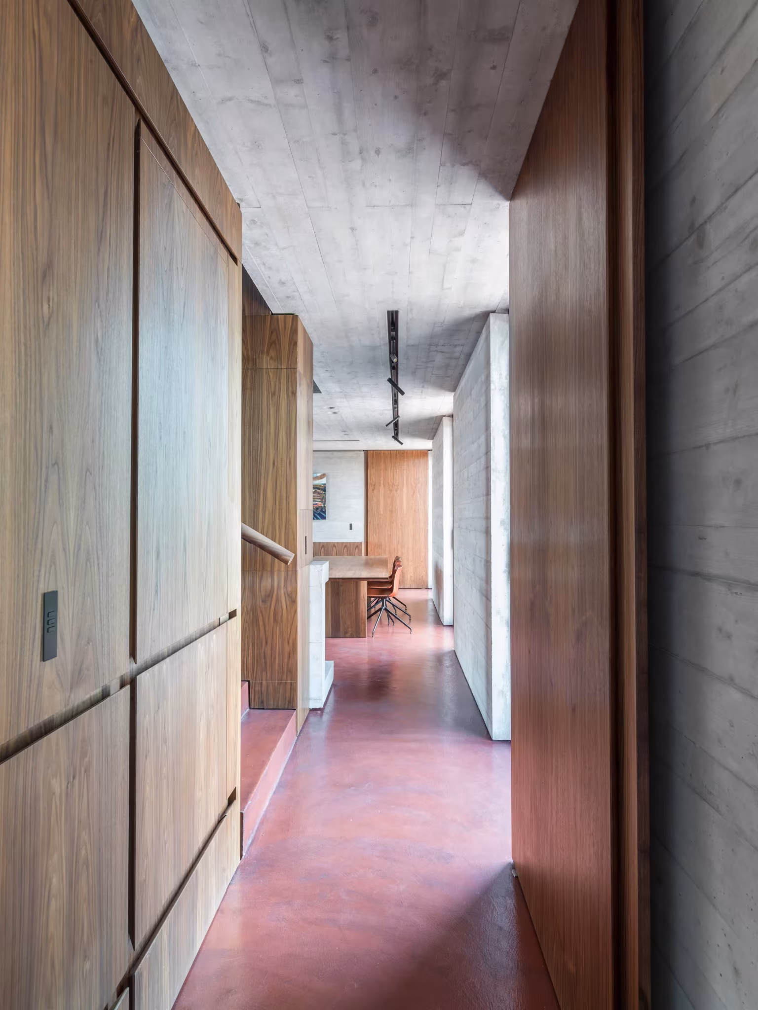 Pasillo entre paneles de madera y hormigón visto con suelo de microcemento rojo en la AP House de Urbino