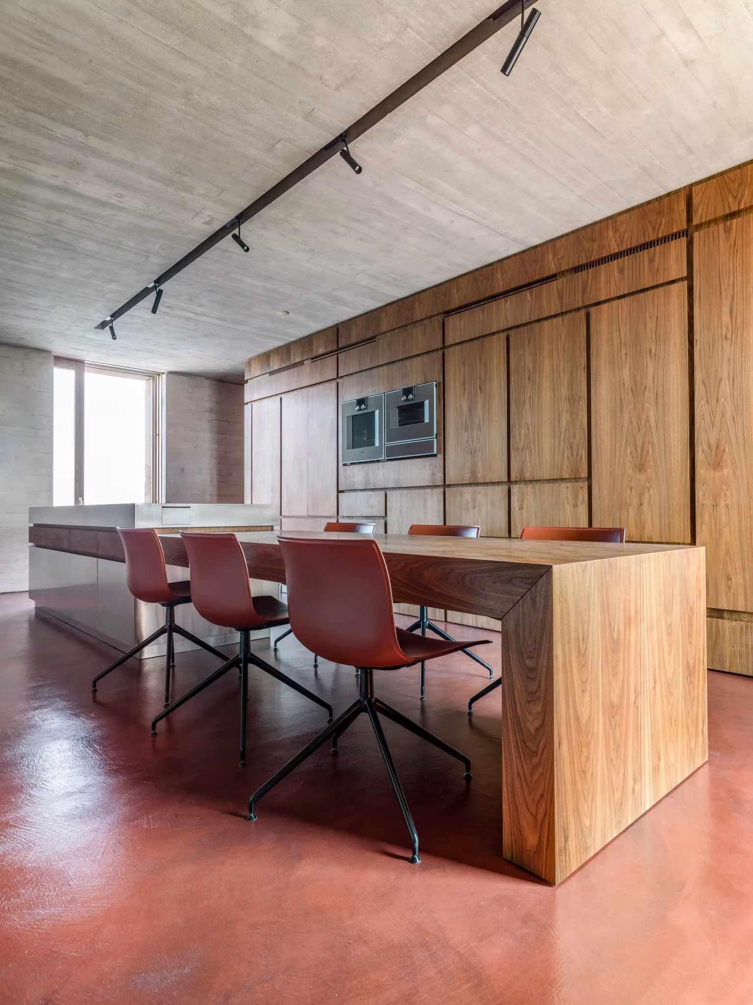 Cocina-comedor con isla de madera, sillas rojas y techo de hormigón encofrado en la AP House de GGA Architects
