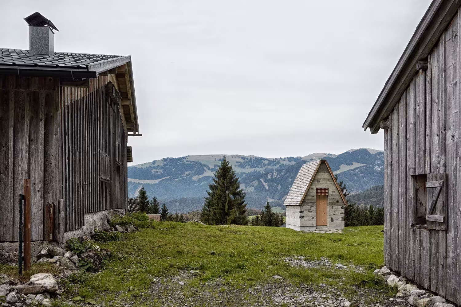 Capilla de Wirmboden entre dos cabañas de madera de la aldea alpina, con montañas al fondo, Innauer-Matt Architekten