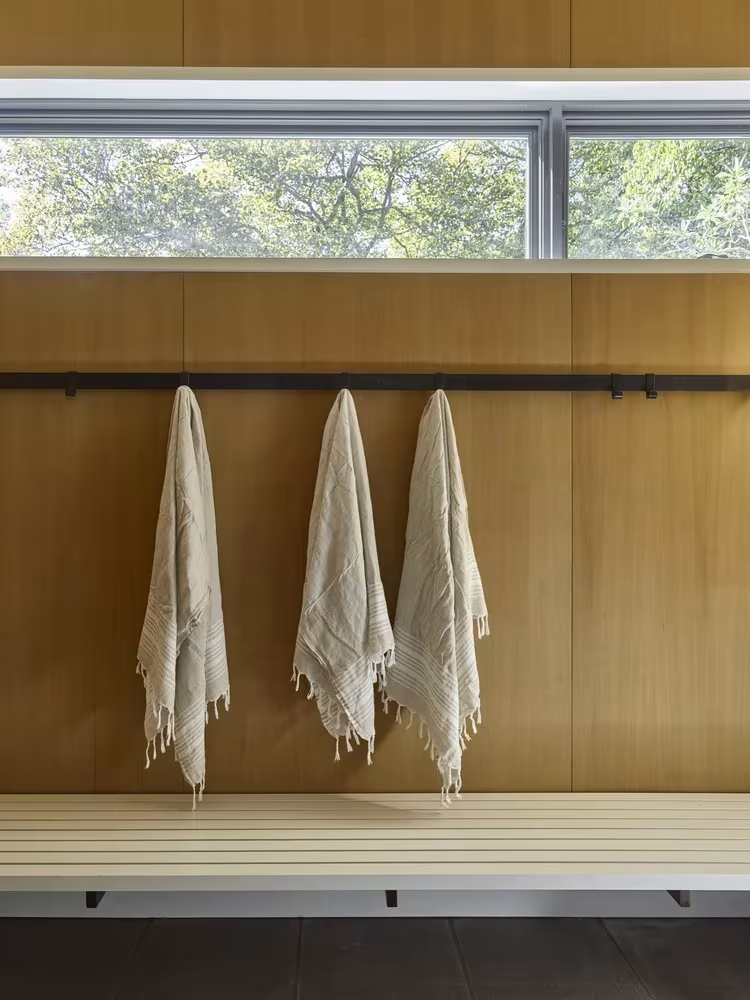 Toallas colgadas de una barra sobre un banco de madera bajo una ventana corrida en la casa de la piscina de Los Hamptons