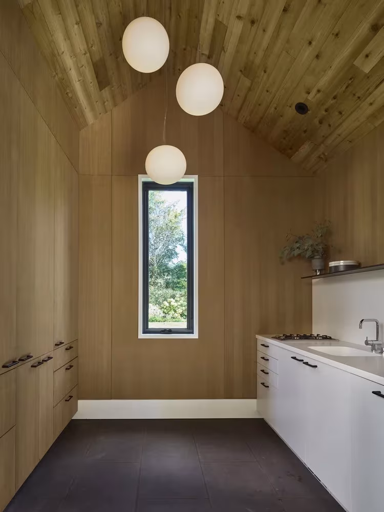 Cocina revestida de madera clara con ventana vertical, lámparas esféricas y techo a dos aguas en la casa de la piscina