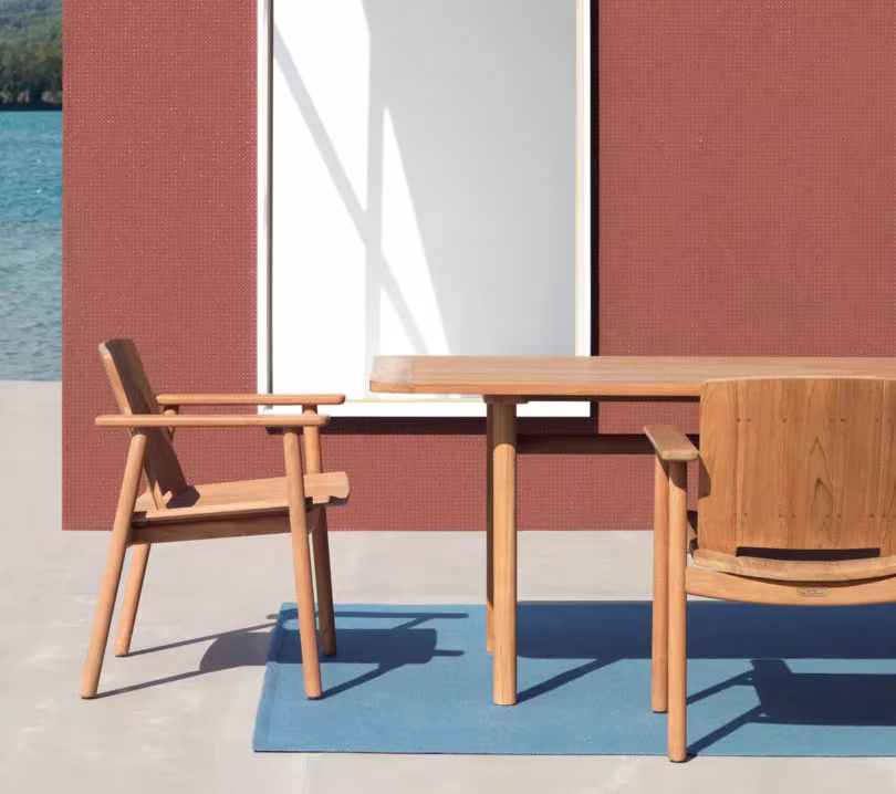 Sillas y mesa de teca de la colección Riva de Jasper Morrison para Kettal sobre alfombra azul ante un muro rojo