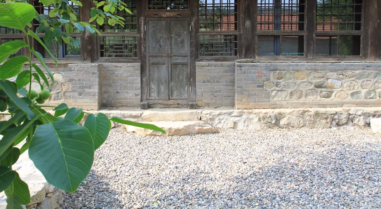 Fachada de la casa rural recuperada por Clou Architects con puerta de madera, zócalo de piedra y grava en primer plano