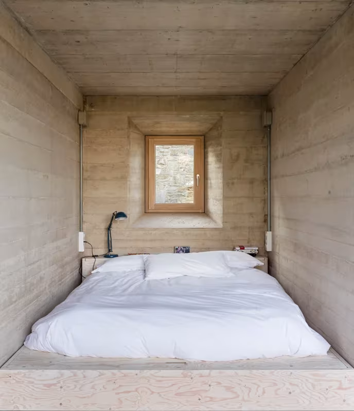 Dormitorio de hormigón encofrado con cama sobre tarima y ventana de madera en la Casa 1413 de Ullastret