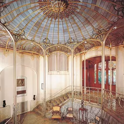 Interior Art Nouveau con cúpula de vidrio sostenida por nervios metálicos curvos sobre una escalera con barandilla