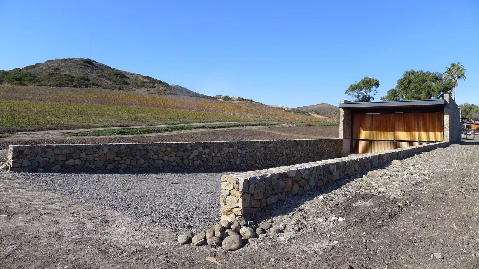 Muro de piedra y pabellón con cubierta oscura en el paisaje árido de Cuatro Cuatros, Ensenada