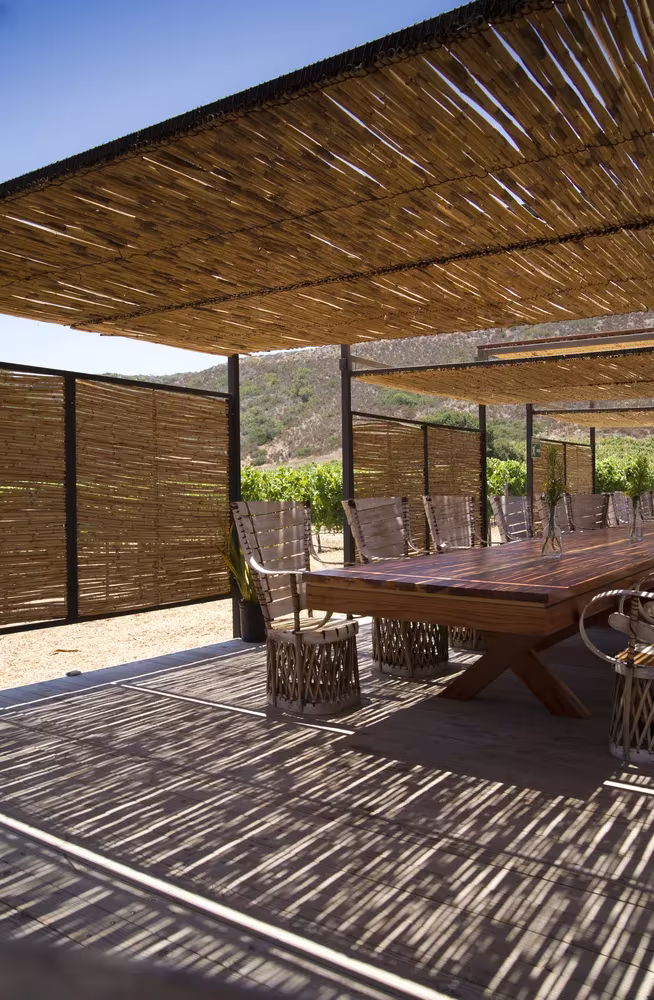 Mesa larga de madera bajo pérgola de cañas y paneles de bambú en Cuatro Cuatros, entre viñedos de Ensenada