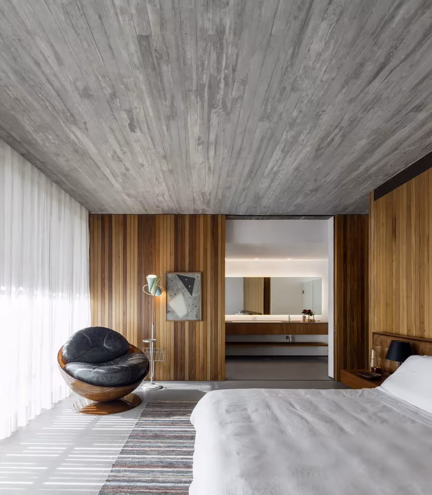 Dormitorio de la Casa plana con techo de hormigón, paneles de madera, cortinas blancas y butaca de cuero, MK27