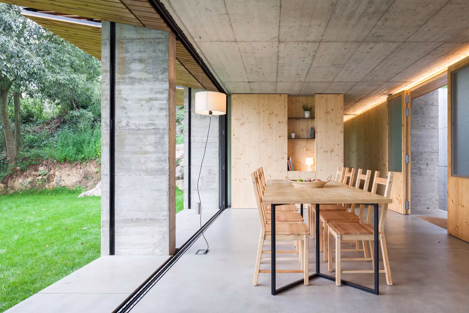 Comedor con mesa de madera, techo de hormigón visto y vidrio corredero sobre el jardín en la Casa Retina de Arnau