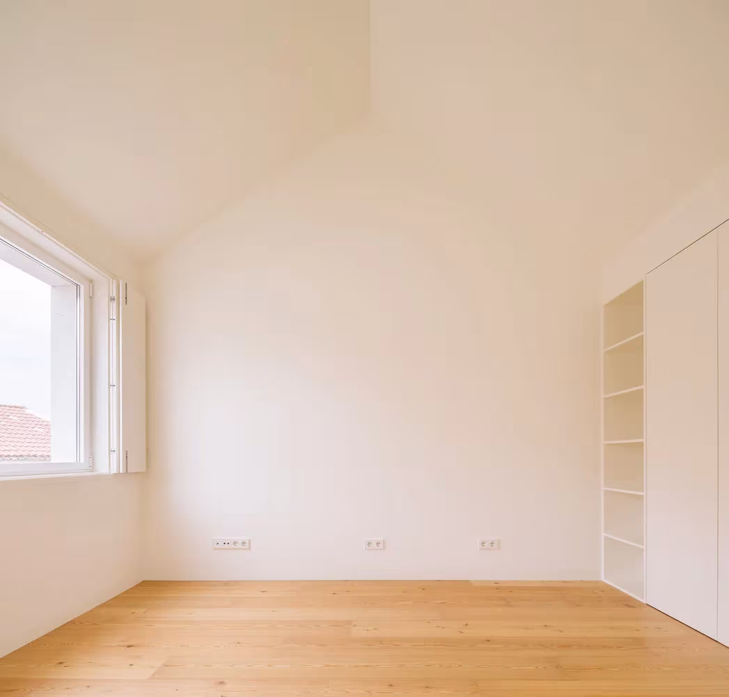 Dormitorio blanco de techo inclinado, ventana de madera y suelo de tarima en la Casa Brotero, Lisboa