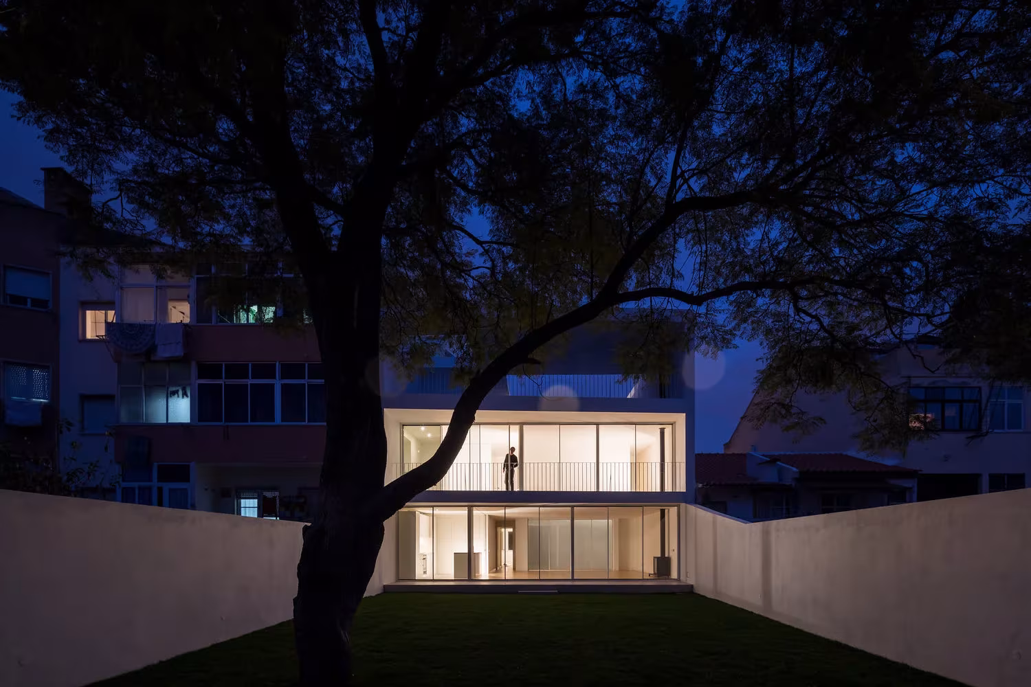 Jardín trasero de la Casa Brotero con muros blancos, césped y árbol, y salón iluminado de noche, Lisboa