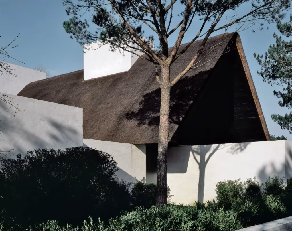 VO Residence de Vincent Van Duysen en Knokke, volumen blanco con cubierta de paja a dos aguas junto a un pino