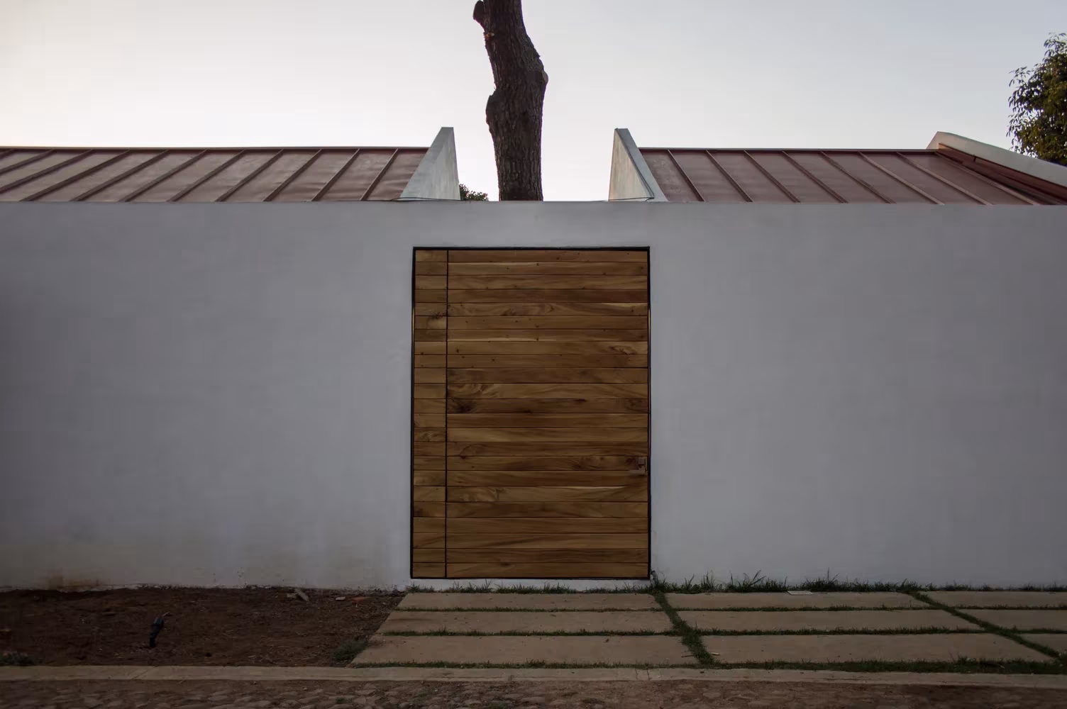 Puerta de madera en el muro blanco de la casa de Espacio EMA con el tronco del pino sobre la cubierta, Jalisco