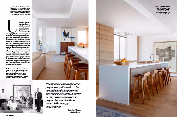 Doble página de INTERIORES con un salón-comedor de suelo de madera y mesa larga ante las ventanas sobre la ciudad