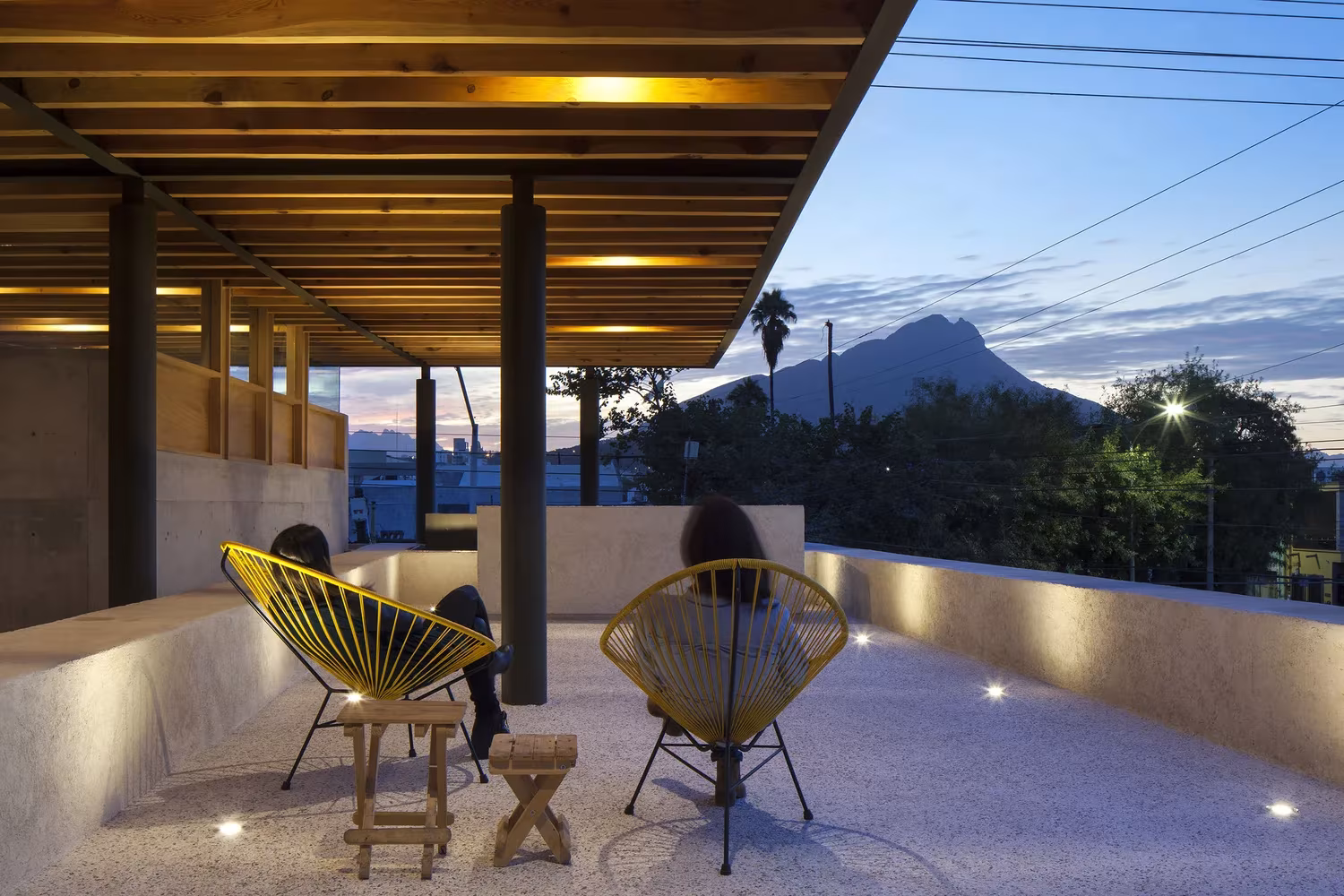 Terraza cubierta con vigas de madera y sillas de colores, con la montaña al fondo, en las oficinas 15 de Mayo de Monterrey