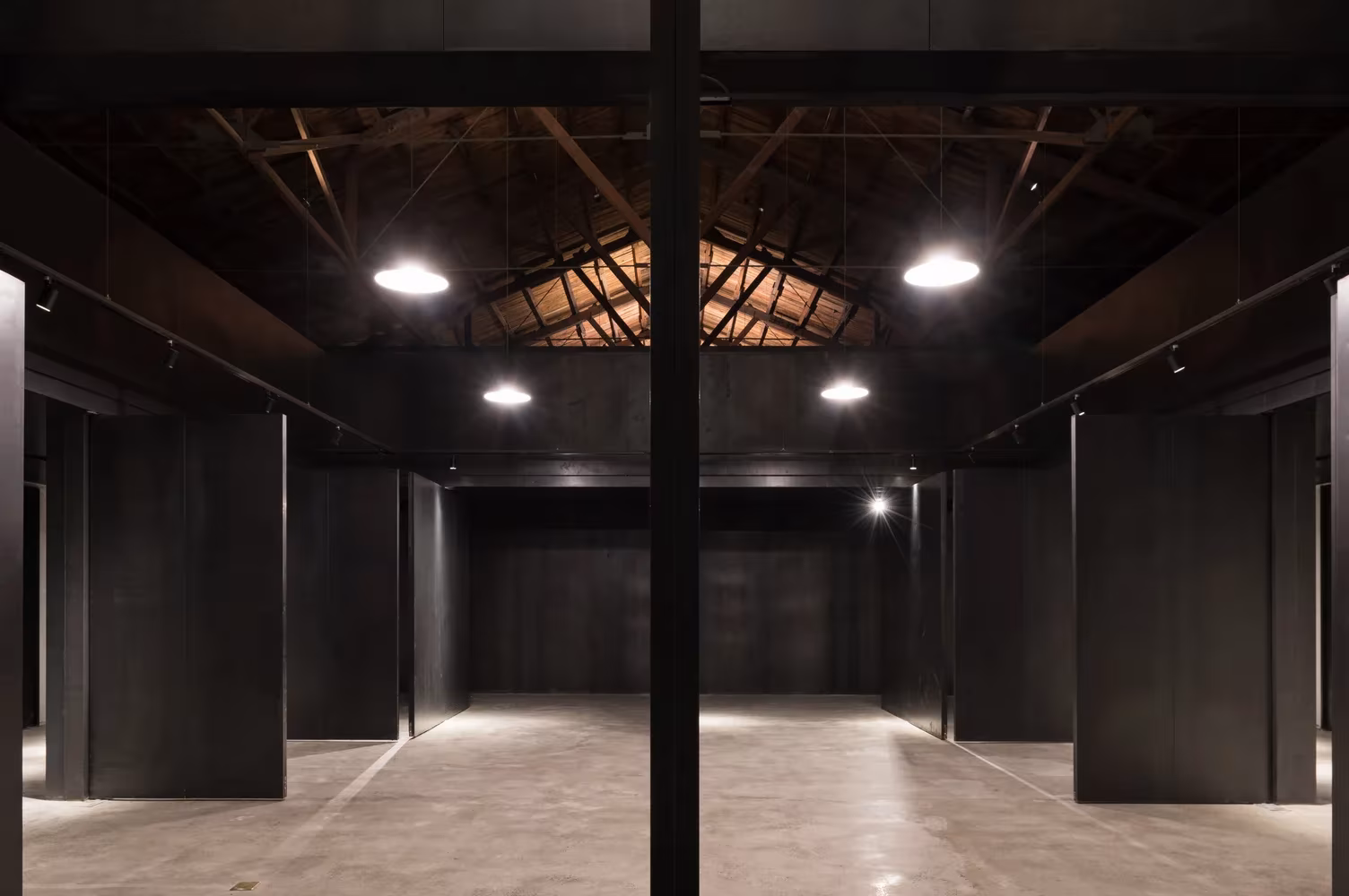 Nave de paneles negros con luminarias circulares y pilar de acero, vista en profundidad, en Lens Magazine de TAO