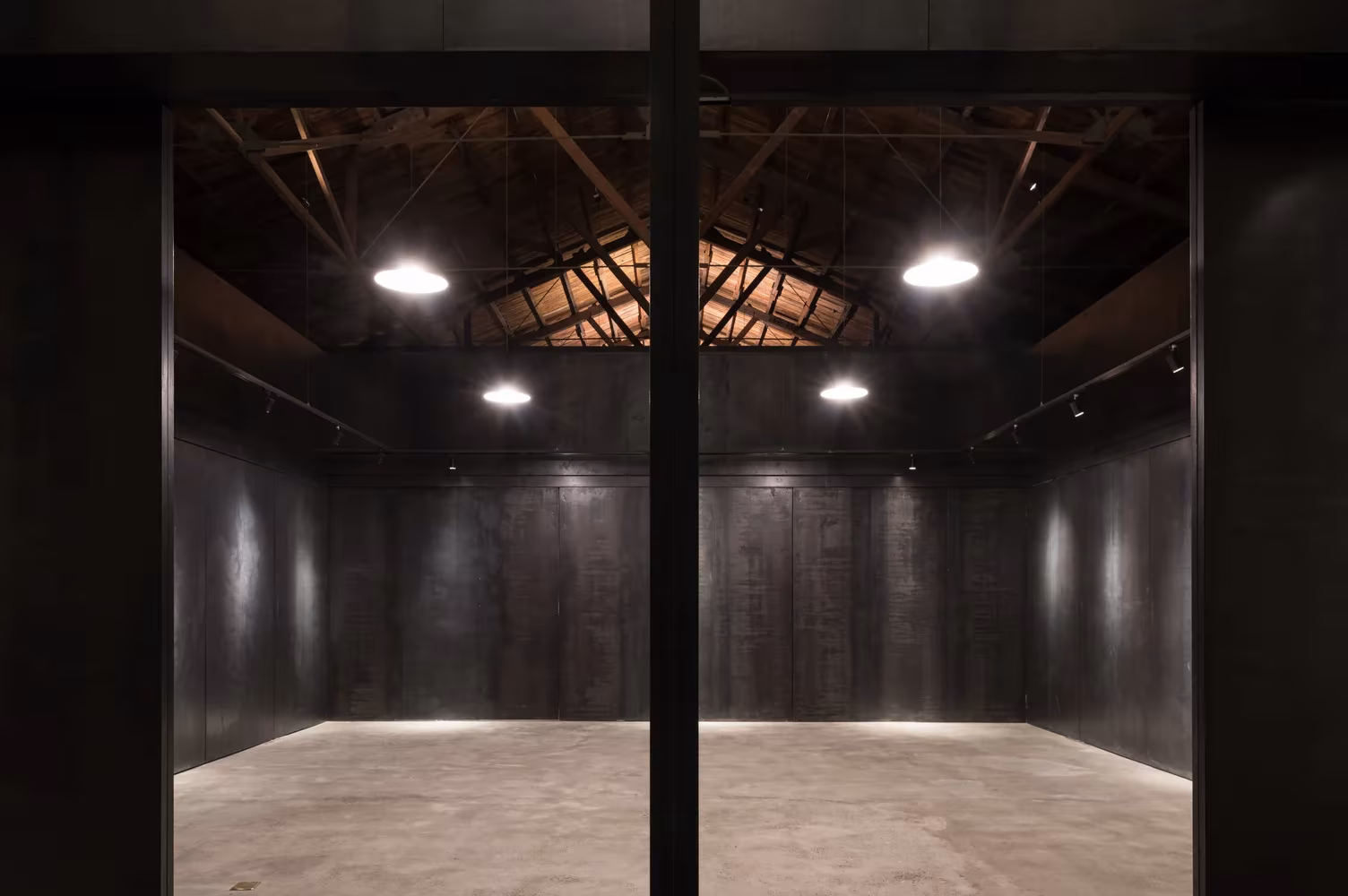 Sala de paneles de acero negro con focos circulares y suelo continuo bajo la cercha del almacén de Pekín