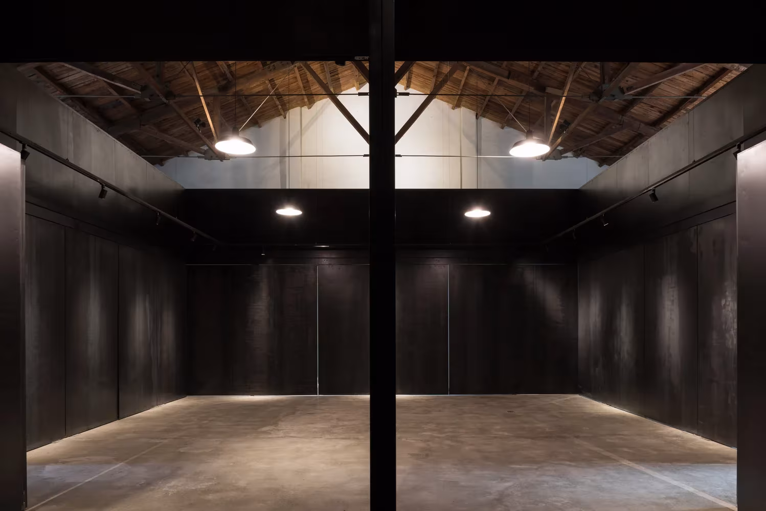Sala diáfana de paneles negros con pilar metálico central bajo la cubierta de cerchas de Lens Magazine, de TAO