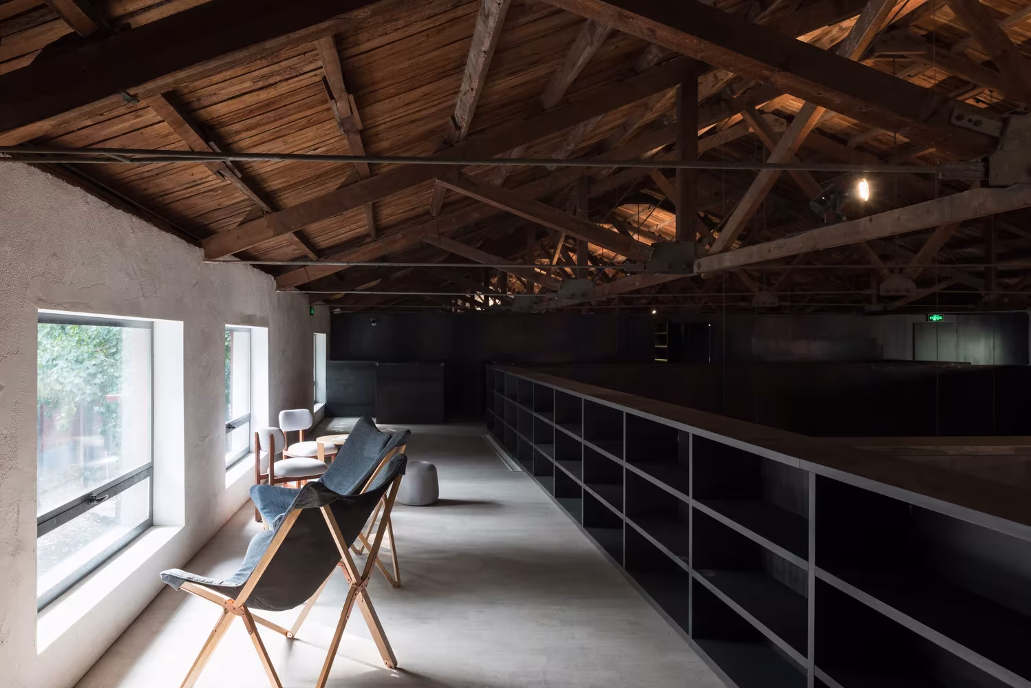 Galería superior con estanterías bajas de acero, sillas de lona y ventana bajo la cubierta de cerchas de Lens Magazine
