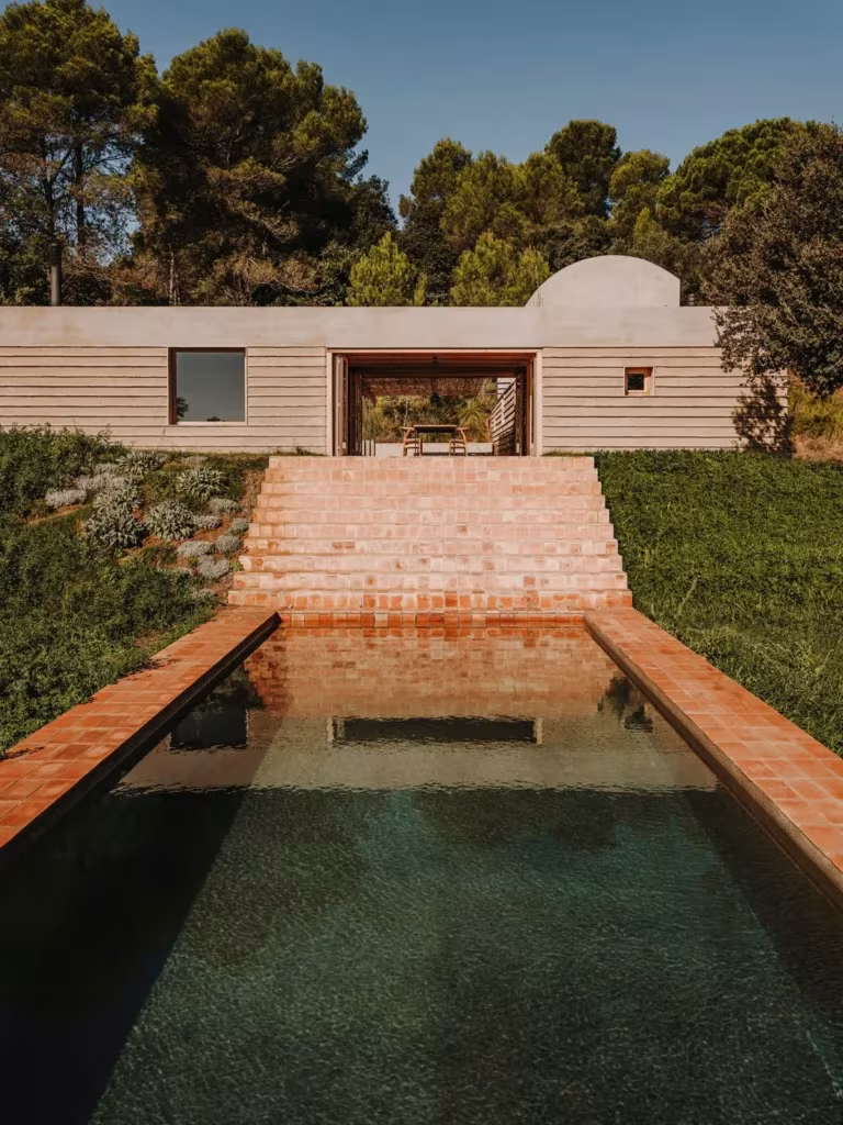 Casa Ter del estudio Mesura, volumen de piedra entre encinas con escalinata de ladrillo y estanque delante