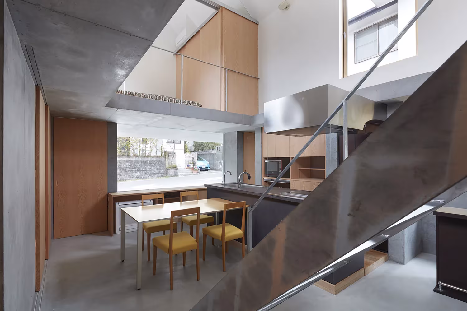 Cocina y comedor a doble altura con mesa de madera y escalera metálica en la casa de Tomohiro Hata en Otsu