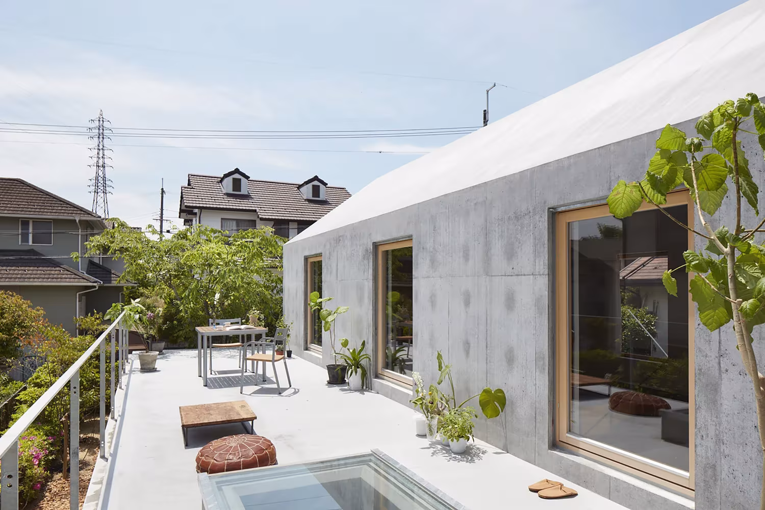 Terraza de hormigón con mesa, pufs y lucernario de vidrio ante la ventana de madera, en la casa de Tomohiro Hata en Otsu