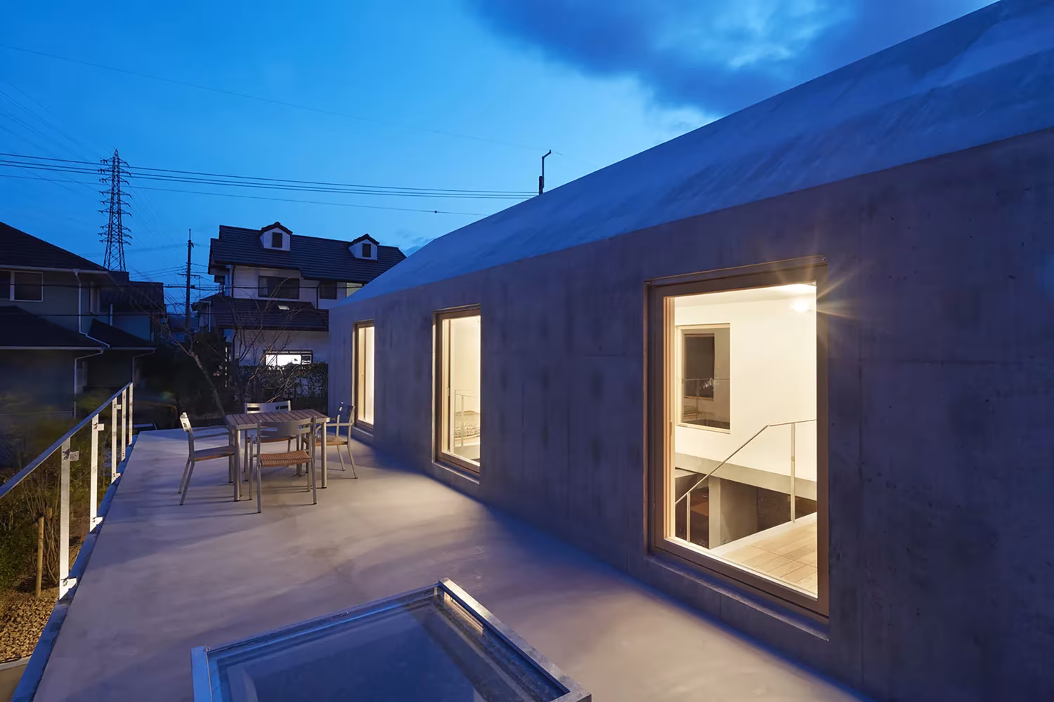 Terraza elevada de hormigón con lucernario de vidrio y ventanas iluminadas al anochecer en la casa de Tomohiro Hata