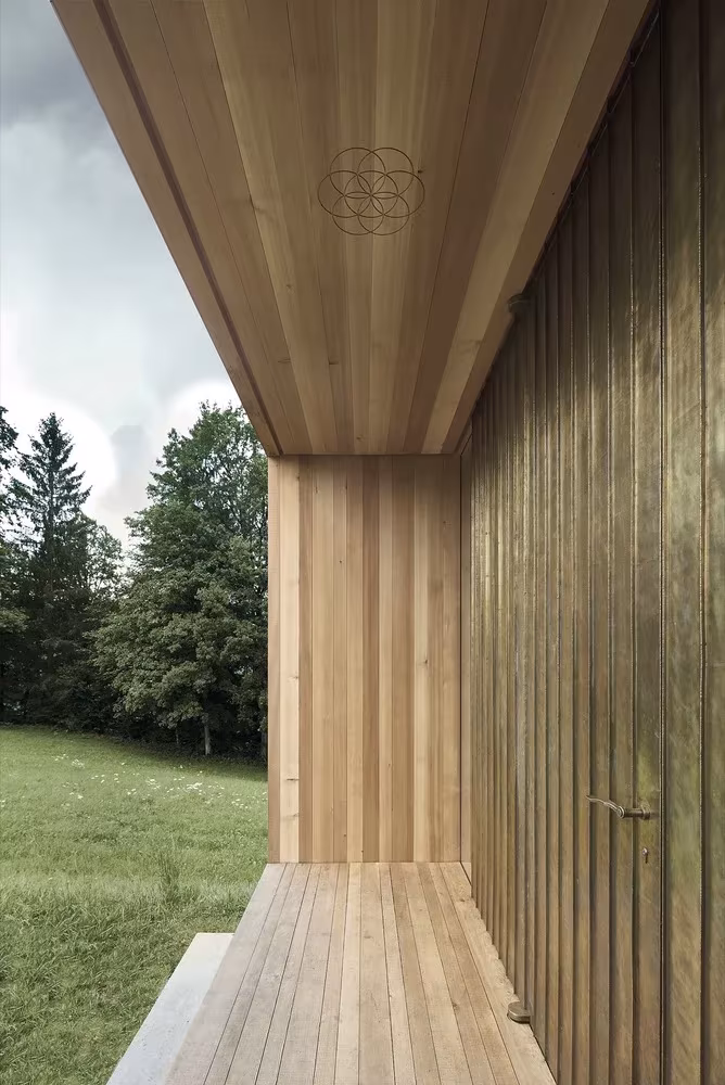 Porche de la Capilla Salgenreute con techo de madera y puerta revestida de latón, de Bernardo Bader