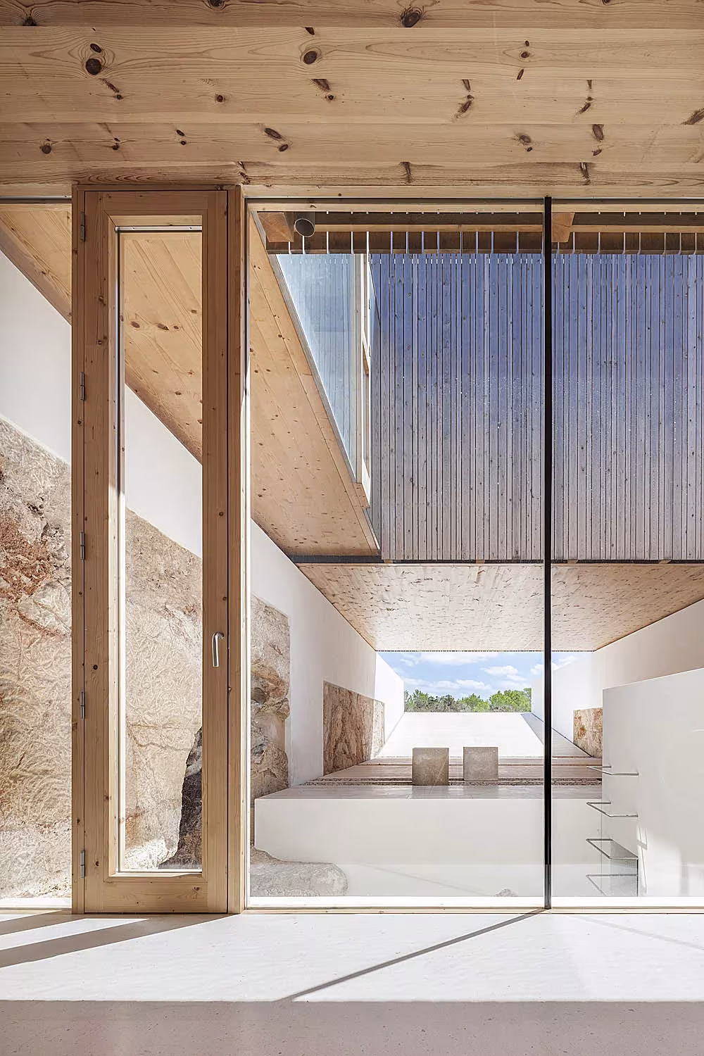 Salón blanco con carpintería de madera abierto a un muro de roca natural en Bosc d'en Pep Ferrer
