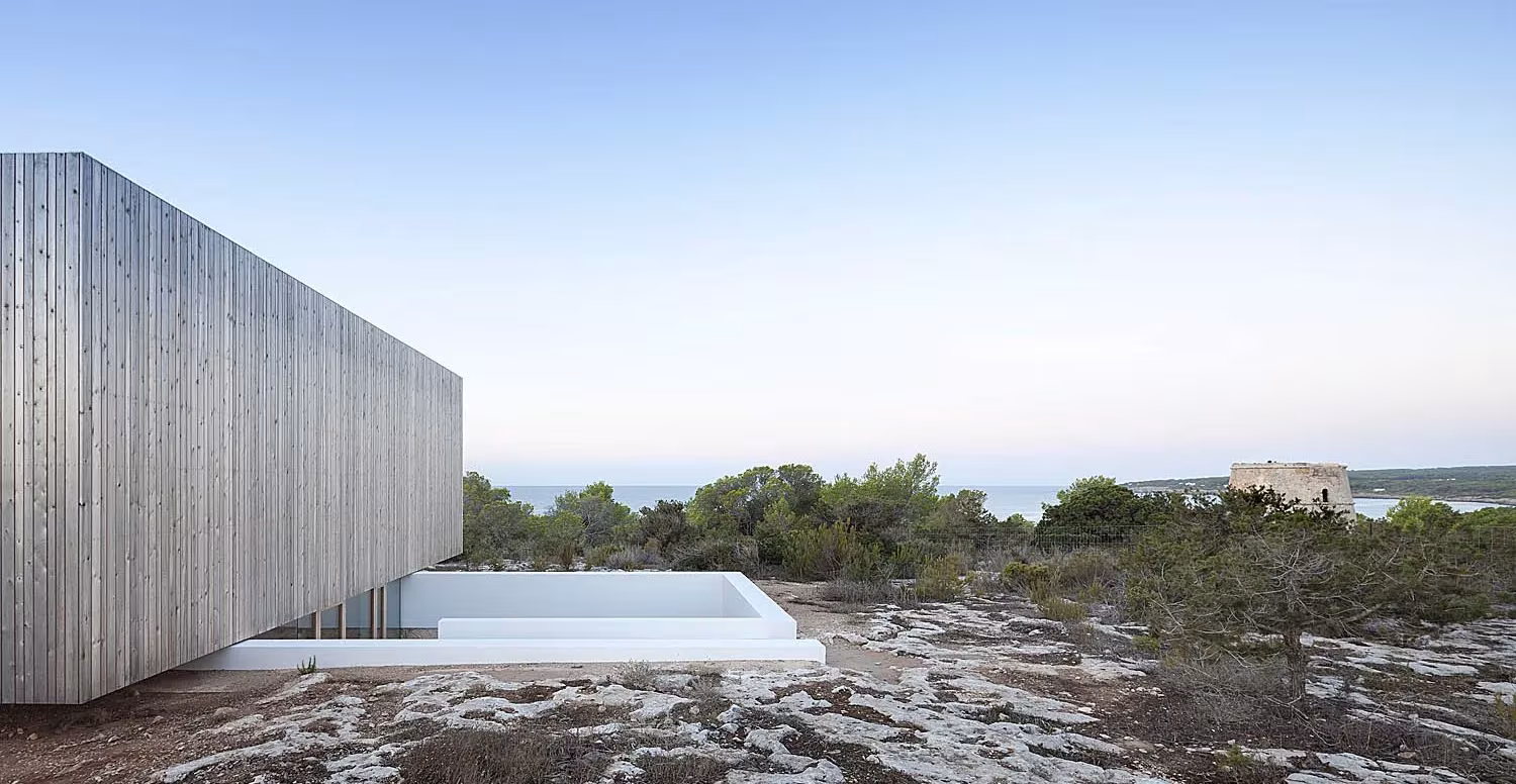 Volumen de madera gris con piscina blanca encastrada en la roca y el mar al fondo, en Bosc d'en Pep Ferrer