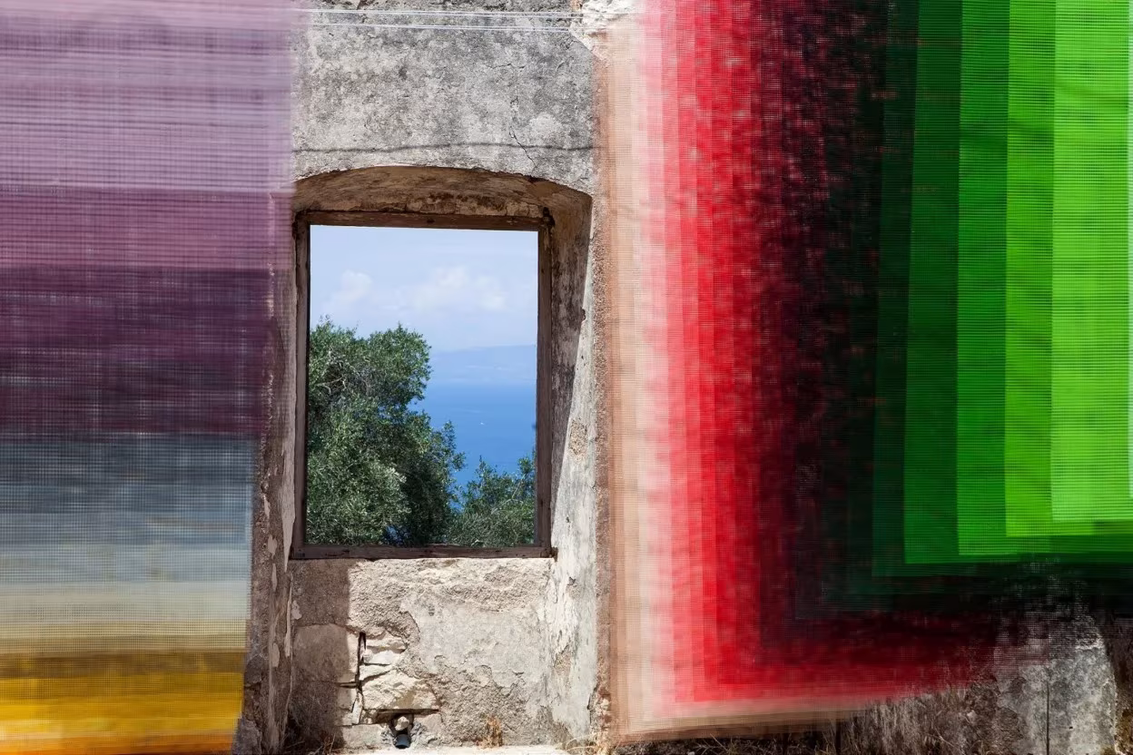 Telas de Quintessenz en verdes, rojos y violetas enmarcando una ventana de piedra con vistas al mar en Paxos