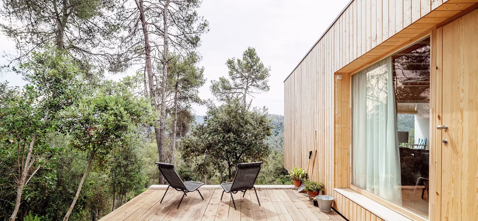 Terraza de madera con tumbonas ante la fachada de listones y el gran ventanal de la Casa LLP en Collserola