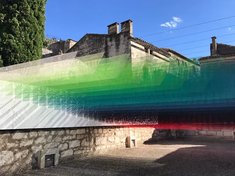 Velo de hilos de colores de Quintessenz suspendido junto a un muro de piedra antiguo bajo cielo azul