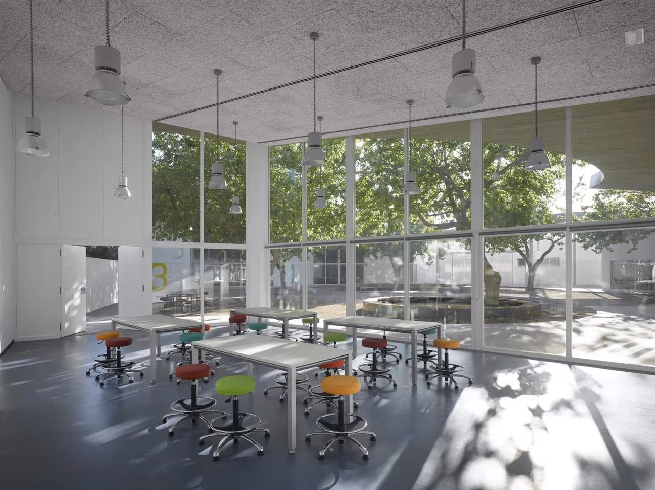 Aula con mesas blancas y taburetes de colores tras un muro cortina arbolado en la Universidad Popular Infantil de Gandía