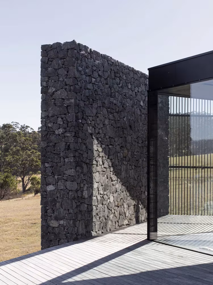 Muro de piedra exento junto a la cristalera y la terraza de madera en D'Entrecasteaux House, de Room11