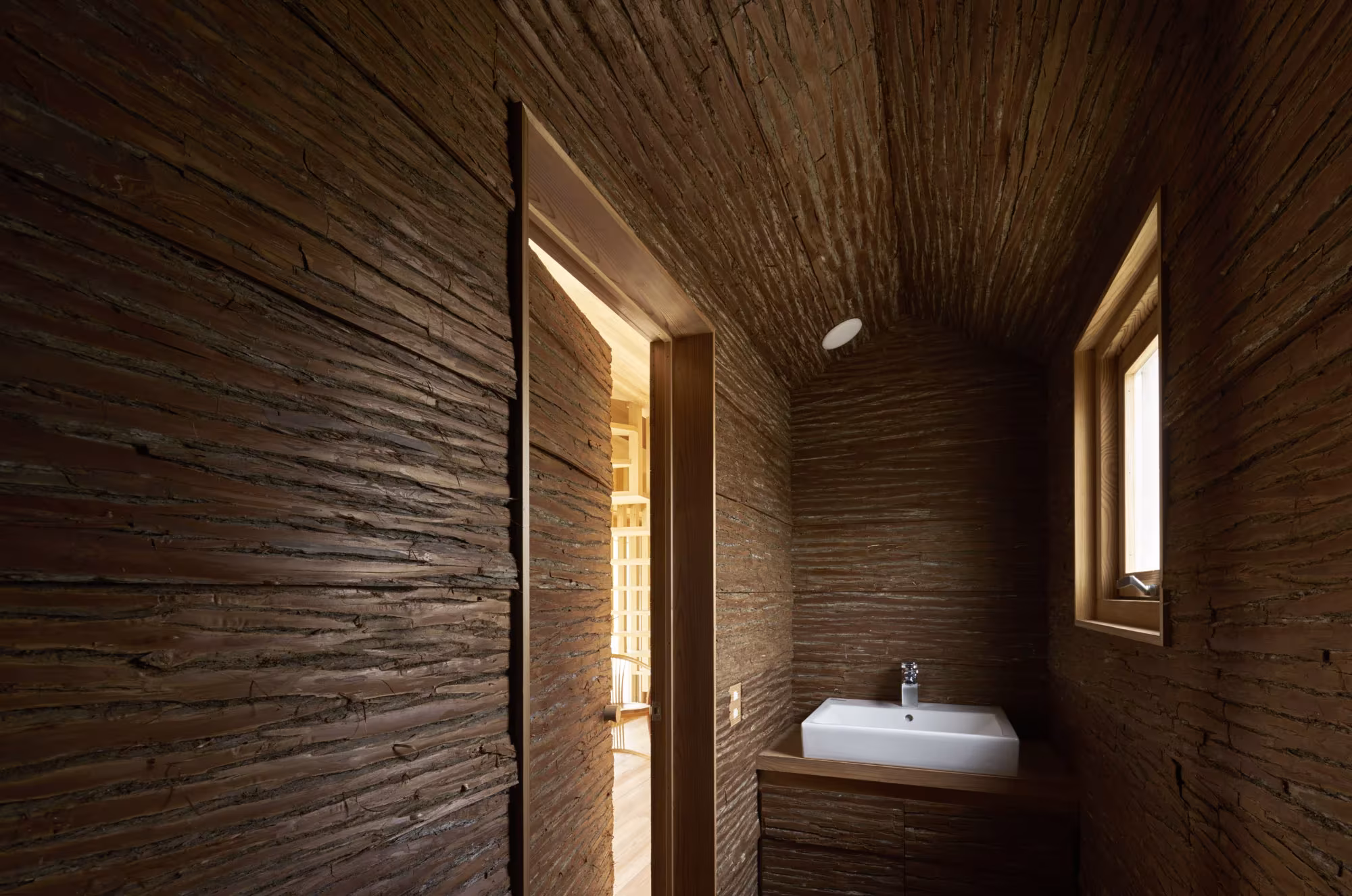 Pequeña estancia de madera oscura con un lavabo blanco, una ventana y una puerta abierta en la Yoshino Cedar House