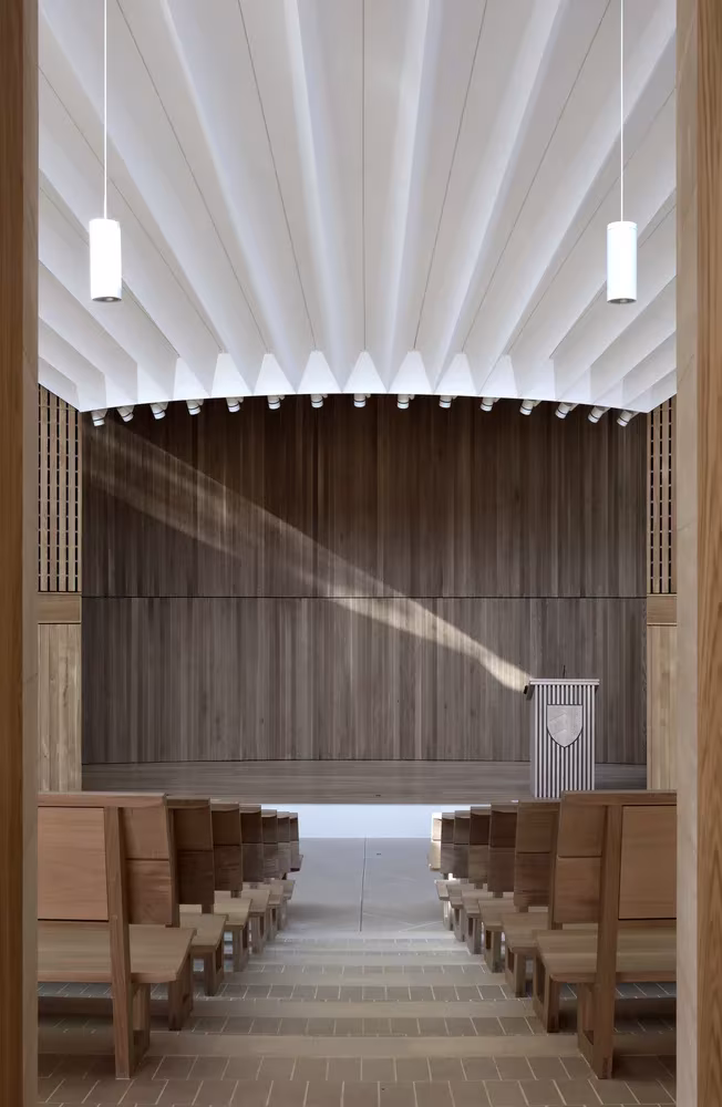 Vista axial del auditorio con pliegues blancos en el techo, panelado de madera y bancos, de Níall McLaughlin