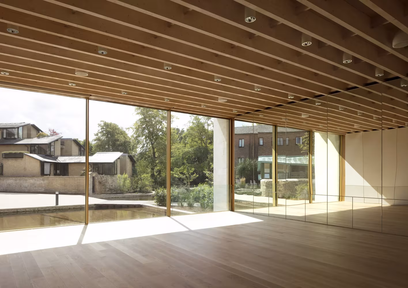 Sala diáfana con vigas de madera y cristalera que da a un estanque y a edificios del campus de Oxford