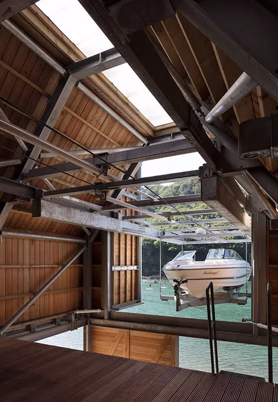 Interior de madera y acero del Berberis Boathouse con una lancha suspendida bajo el lucernario, en Devon