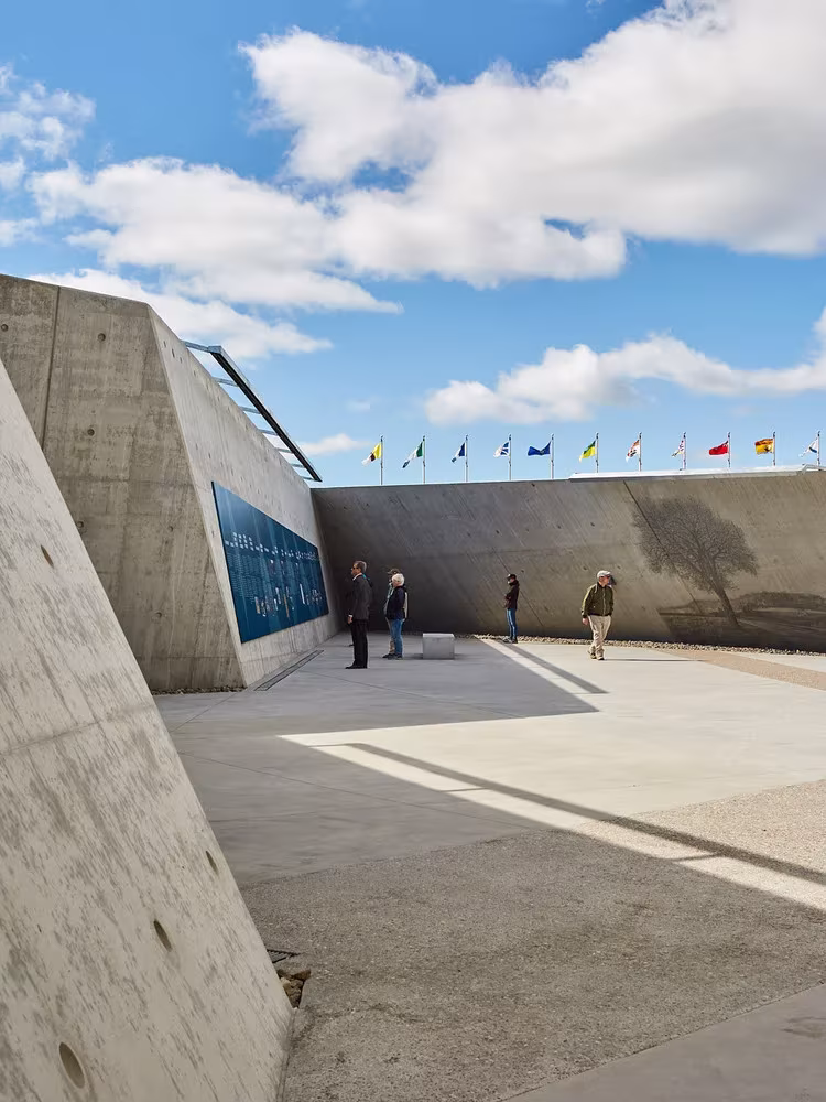 Visitantes junto a un muro de hormigón inclinado con banderas al fondo en el Monumento del Holocausto de Ottawa