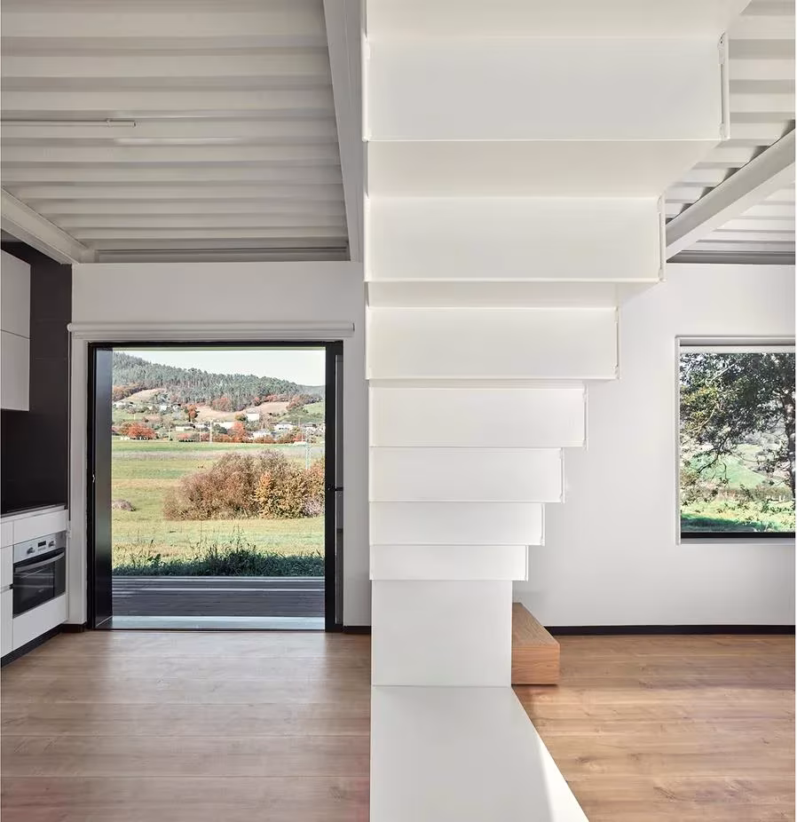 Interior blanco con escalera de peldaños volados y ventanal con vistas al valle en la Casa Montaña de Baragaño