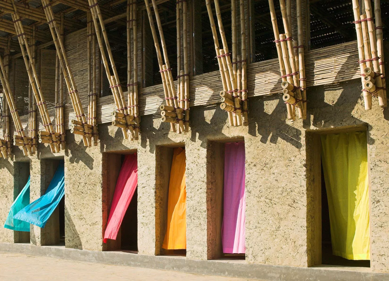 Muro de barro y paja con vanos de cortinas de colores bajo estructura de bambú en Bangladesh, de Anna Heringer