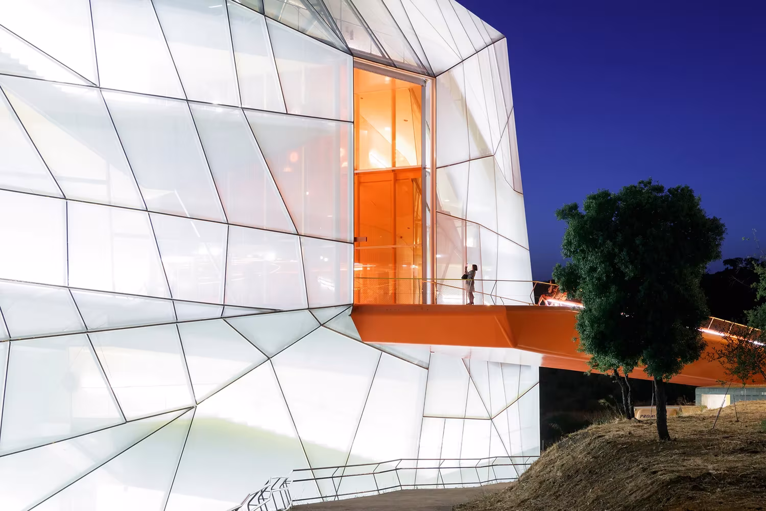 Fachada translúcida de nervios ligeros y pasarela naranja del Palacio de Congresos de Plasencia al anochecer
