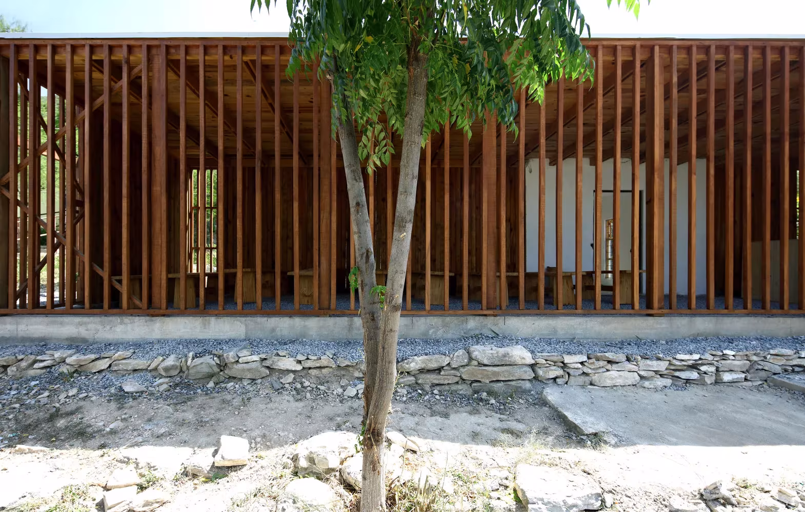 Cerramiento de listones verticales de madera sobre un zócalo de piedra y un árbol en la Parroquia de San Rafael Arcángel