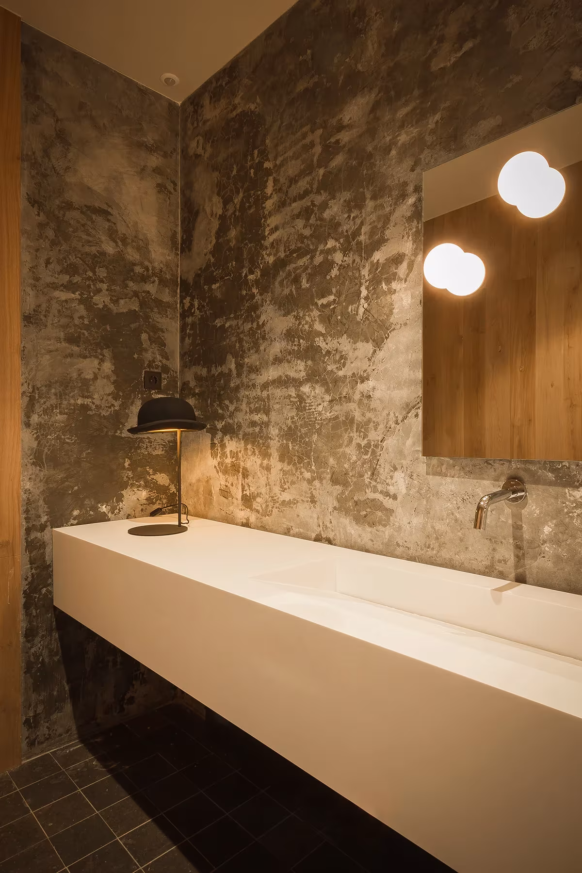 Lavabo corrido blanco ante un muro de piedra desnuda con espejo y apliques redondos en Sant Sadurní de Callús
