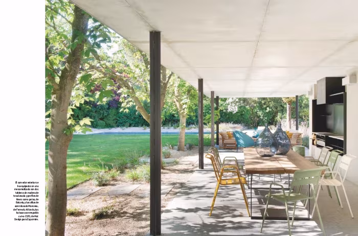 Porche de la casa de ÁBATON con pilares metálicos, mesa de madera y sillas de colores abierto a un jardín de césped