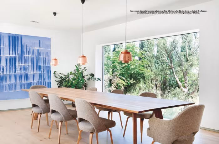 Comedor de mesa alargada de madera, sillas tapizadas, lámparas cobrizas y un cuadro azul ante un ventanal sobre el jardín