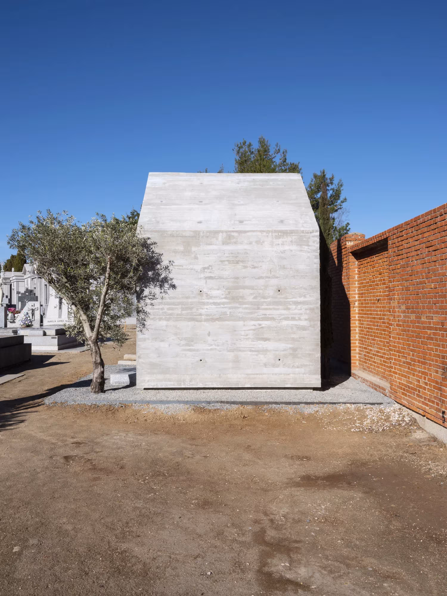Panteón de ÁBATON de hormigón visto con forma de casa, entre un olivo y un muro de ladrillo del cementerio