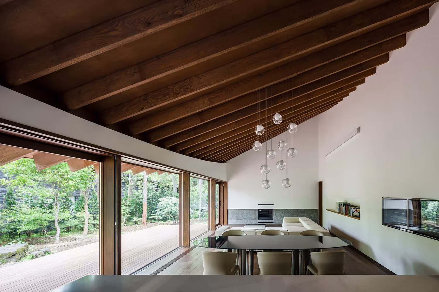 Comedor y cocina bajo la cubierta de vigas de madera inclinadas con ventanales al bosque en Karuizawa