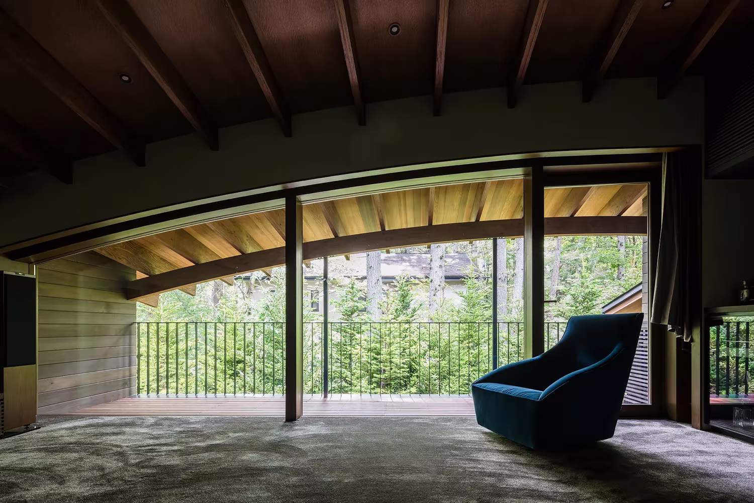 Sala bajo la cubierta curva de vigas de madera con butaca azul y vistas al bosque en Karuizawa